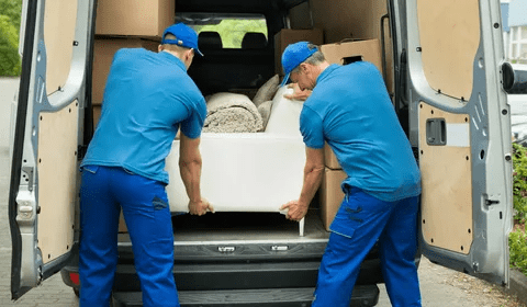 alt= Two movers in blue overalls load boxes and a sofa into a white van on a sunny day, showcasing efficient sofa removal in London.